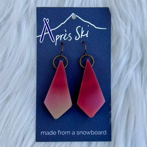 Pink teardrop snowboard earrings on a blue card with 'Apres Ski' branding, against a white fur background.
