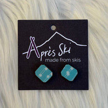 Teal geometric earrings on a black card with 'Apres Ski' branding, against a fur background.