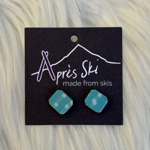 Teal geometric earrings on a black card with 'Apres Ski' branding, against a fur background.